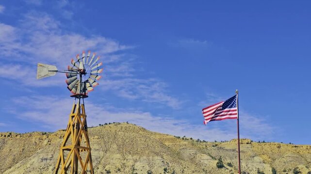 Old fashioned wind mill turning in the wind with American flag waving along side against blue sky with light clouds and buff collared hills and cliffs