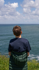 Fototapeta premium Teenage boy standing on coastal cliff and looking at ocean horizon in Asturias, Spain. Peaceful travel and nature scene with Atlantic sea view and dramatic cloudy sky.