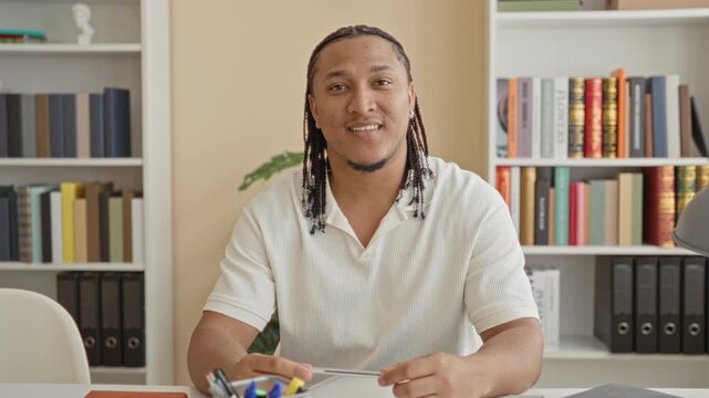 Man wearing white polo and braids, holding a pen over an open notebook, hands on paper at a study desk with bookshelves, binders and a potted plant behind him; focused learning.