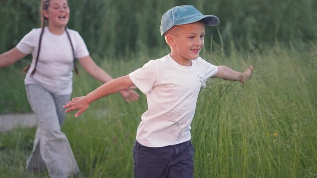Boy running outdoors open arms. Girl follows boy in grass. Child enjoying outdoors freedom. Boy plays fun running with arms wide. Girl runs after boy in outdoors park. Children love freedom and fun.