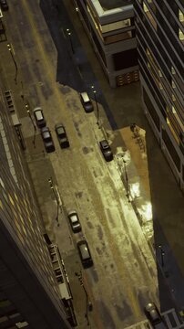 People are walking on the street near a tall building at night. City lights are shining on the sidewalk. Cars are parked along the road. Windows are lit in the building above.