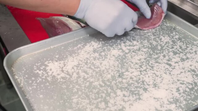 High-angle shot of a chef's hands in blue gloves preparing fresh fish fillets by generously coating them with sea salt on a large metal baking sheet. This professional culinary process is essential fo