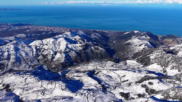 Cinematic drone reveal shot flying over snow-covered winter hills to uncover a beautiful coastal city by the deep blue sea. Bright sunny day with vibrant blue sky stretching to the horizon. Perfect tr