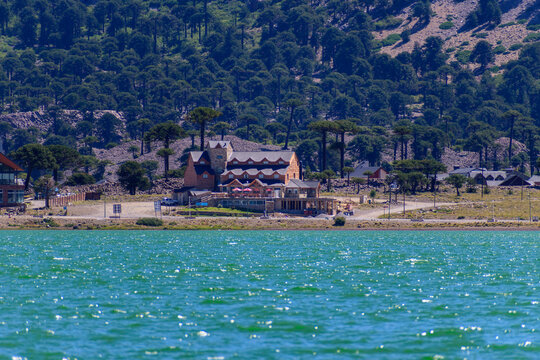 Hotel de monta&ntilde;a frente a un lago de aguas turquesas y rodeado de un bosque alpino de araucarias en un d&iacute;a soleado de verano.