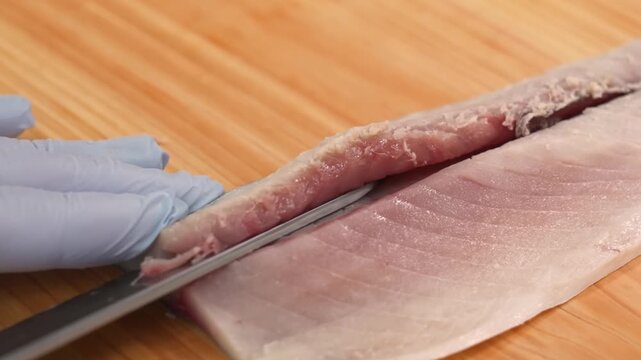 High-quality footage of a professional chef or cook wearing blue gloves skillfully using a sharp knife to fillet fresh raw fish on a rustic wooden cutting board. This detailed shot captures the culina