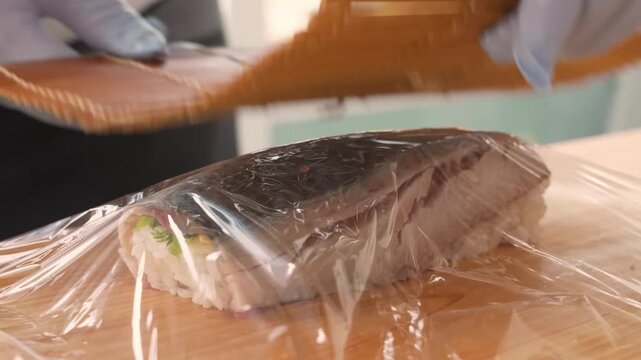 A detailed, slow-motion shot showing a professional chef using clear plastic wrap to shape and tighten a fresh sushi roll on a wooden cutting board. The scene highlights the intricate process of prepa
