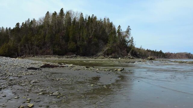 Nature drone footage documenting the powerful outflow of the Mitis River into the St. Lawrence estuary beneath an overcast sky in Gasp&eacute;sie, Quebec.