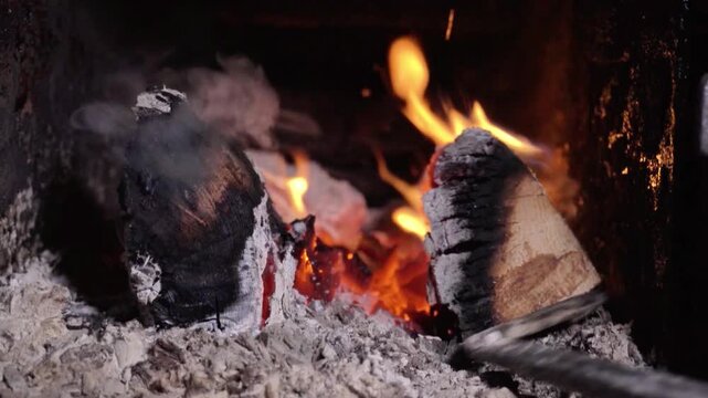 Flames flicker as a metal rod stirs glowing embers and logs in a fireplace, showcasing the warmth and light of a cozy fire setting