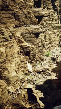 The scene shows large rock formations next to a body of water in a canyon. The sun shines on the rocks, revealing their texture and color. Plants grow along the edge of the water.