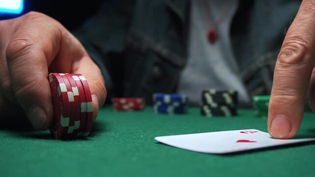 Player places a red poker chips on a green felt table, pointing at a playing card featuring a heart symbol, with various poker chips arranged in the background under dim lighting