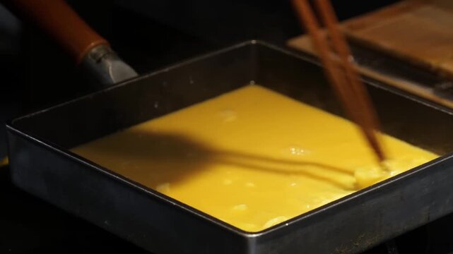 Close-up footage of a chef expertly cooking Tamagoyaki, a traditional Japanese rolled omelette, in a specialized rectangular tamagoyaki-ki pan. The video captures the process of pouring and rolling th