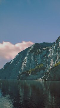 A rocky shoreline sits beside still water under a clear sky. The sun shines on the cliffs, reflecting on the water as clouds drift above. This scene shows natures beauty in a coastal area.