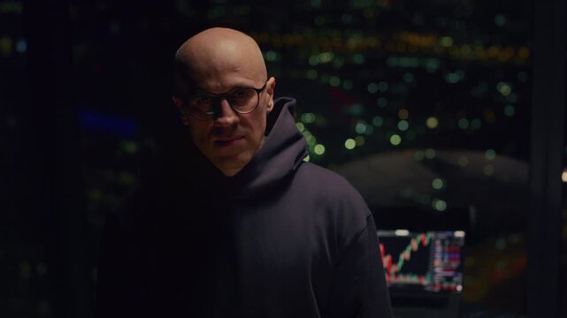 Portrait: bald man trader trades on stock exchange on laptop at home at night, lives in a skyscraper, night city. Male investor analyzes stock market and cryptocurrencies. Apartment in downtown. BTC
