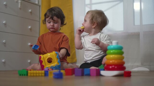Two young toddlers sit on bedroom floor playing with colorful educational toys. Children explore bright building blocks, stacking rings and shape sorters during interactive playtime together.