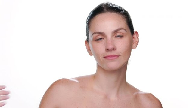 Portrait of fair-skinned brunette Latina with freckles with blue eyes and ponytale hairstyle strokes clavicles turning her body to the camera presenting balanced skin against white bg