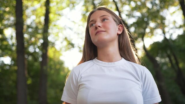 Teenage girl looking up peacefully in forest with sunlight through trees. Teen smiles in nature during golden hour. Young girl breathes fresh air outdoors. Teenager enjoys peaceful forest.