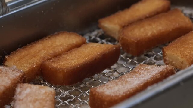 close-up shot of crispy, deep-fried bread sticks generously coated in cinnamon sugar, resting on a metal cooling rack in a professional kitchen or bakery setting.