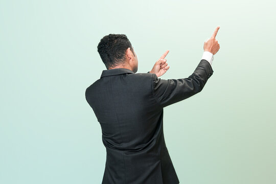 Rear view of an Indonesian professional man in a black suit pointing both index fingers toward the upper right corner against a green background.
