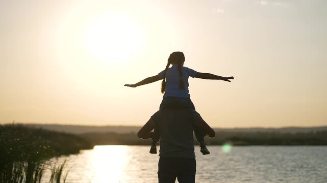 Father carries daughter on shoulders at sunset. Child spreads arms wide by lake. Dad supporting kid outdoors. Family enjoying golden hour near water. Parent and child silhouette at sunset.