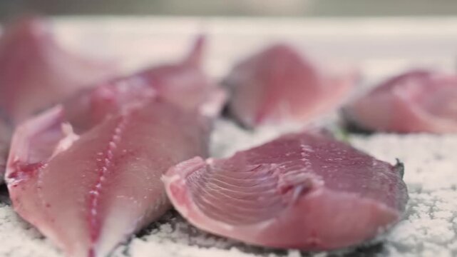 A close-up shot of fresh, pink raw fish fillets being seasoned with coarse salt on a stainless steel tray. This footage captures the initial steps of a culinary preparation, highlighting the texture a