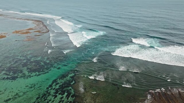 Ocean waves are breaking over a vibrant turquoise coral reef and shallow waters on Bali's Bukit Peninsula, creating a dynamic natural pattern. Nature background. Aerial drone flight panorama