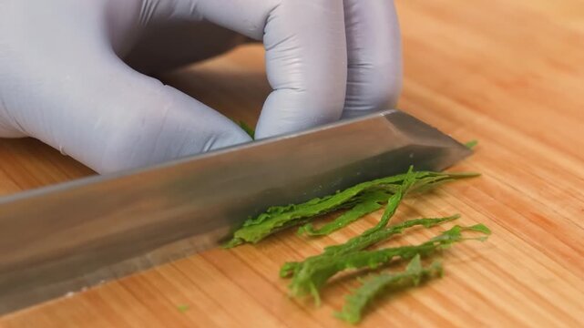 High-angle, close-up footage of a person's hand wearing a protective grey cut-resistant glove while finely slicing fresh green leafy herbs (such as basil, shiso, or spinach) using a sharp professional