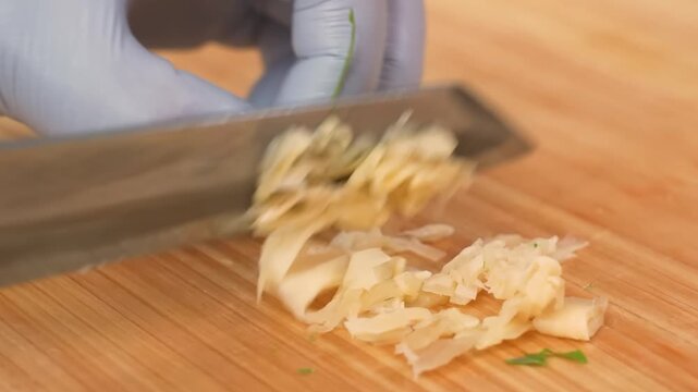 A close-up, high-angle shot of a chef's hands wearing blue nitrile gloves skillfully mincing and chopping fresh garlic cloves with a sharp kitchen knife on a rustic wooden cutting board. The footage h