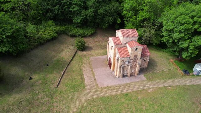 A cinematic drone zoom-out reveals San Miguel de Lillo in Oviedo, a UNESCO World Heritage site, rising above the trees and showcasing its Asturian pre-Romanesque beauty in a serene landscape.
