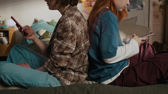 Side view shot of two Caucasian teenage girls sitting back to back on bed while using smartphones in cozy bedroom
