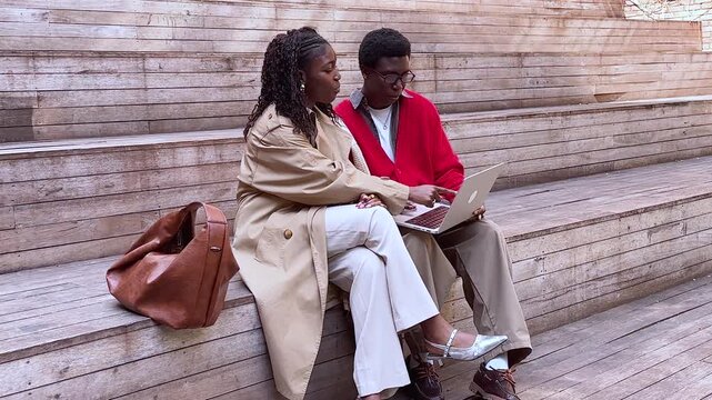 Cheerful african american couple friends man and woman sitting watching media on laptop outdoors in lounge area, smiling talkative black male and female buddies lively discussing video and joking.