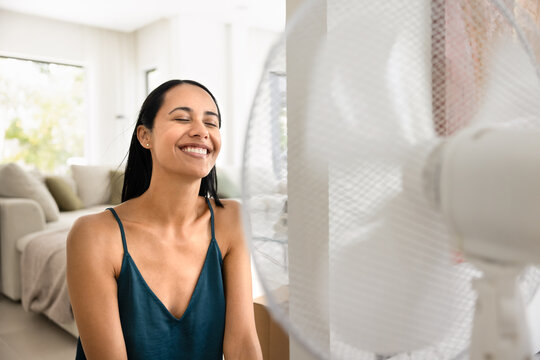 Happy young woman sitting indoors with eyes closed, smiling, feels refreshing breeze from electric fan inside modern living room, enjoying comfort, relief and relaxation during hot summer day at home