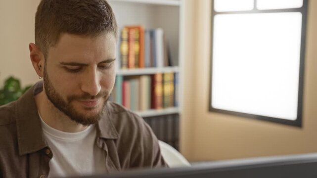 Man smiling at computer monitor, head turned toward screen in a building study with bookshelf, window and plant visible; remote work concentration.