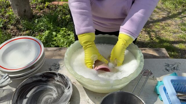 Woman in yellow gloves washing dishes in basin outdoors person scrubs large wooden cutting board with sponge under a kitchen faucet. Bright lighting, close-up shot, steady camera, clean environment