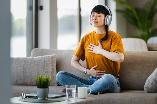 Woman practicing mindfulness meditation with headphones at home on sofa, breathing deeply