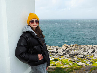 Young woman wears bright yellow beanie and black puffer jacket. She stands near coastal white structure with metal gate. Glasses frame her calm, thoughtful gaze over the sea © mark_gusev