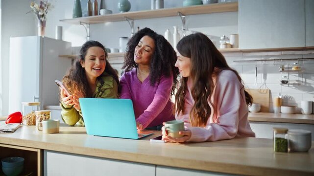 Diverse women ordering online purchases at kitchen using laptop holding phone.