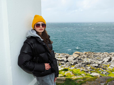 Young woman wears bright yellow beanie and black puffer jacket. She stands near coastal white structure with metal gate. Glasses frame her calm, thoughtful gaze over the sea