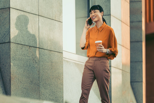 Young professional woman talking on phone and holding coffee outside modern office building