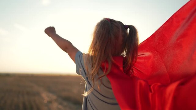 Child wearing red superhero cape runs through sunset field. Young girl with pigtails raises power fist. Kid plays hero with flowing red cape. Childhood imagination outdoors at golden sunset.