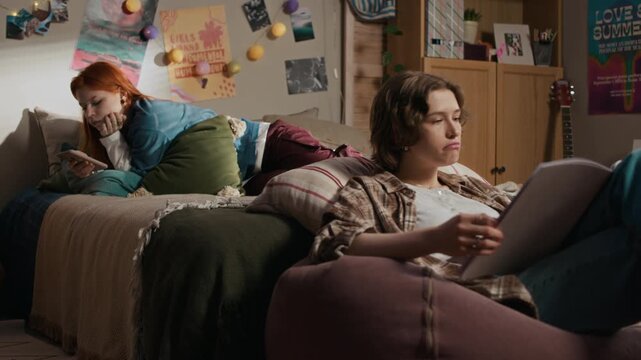 Eye level shot of two Caucasian teenage girls relaxing together in warm bedroom atmosphere, one using smartphone while the other reading magazine on beanbag