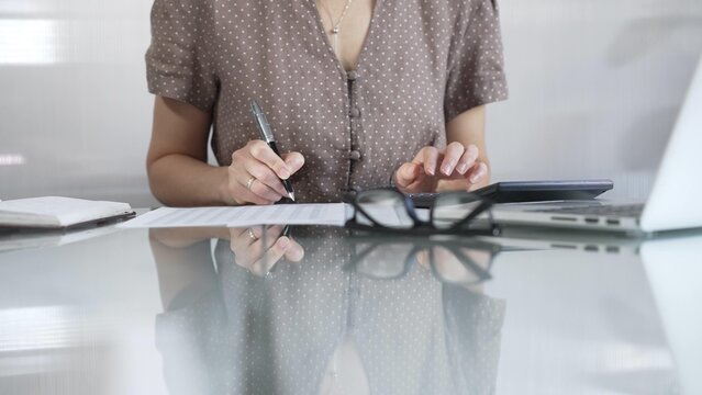 Businesswoman accountant, wearing brawn dotted blouse, is using a calculator to crunch numbers and jotting down meticulous notes, at sleek glass desk in a contemporary office. Taxes, audit in business