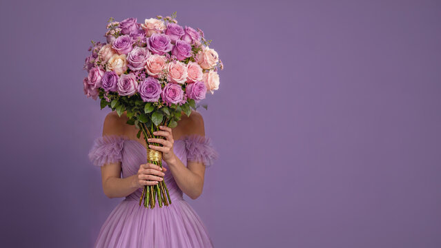 Maid of Honor in a lavender tulle dress holding a large rose bouquet over her face against a purple studio background with ample copy space.