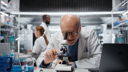Fototapeta premium Smiling senior biologist in medical laboratory using scientific microscope for cellular analysis. Jolly elderly man examining sample in lab with high tech optical device gear, camera A