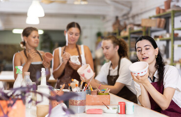 Obraz premium Young woman paints white ceramic blank, vase bowl, decorates product, completes work, makes unique piece of art. During work, girl talks sweetly with friend, communicates, jokes and laughs