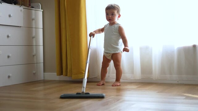 Cute toddler baby in gray tank top and diaper holds a mop on a wood parquet floor. White dresser and bright yellow curtains in background. Barefoot child imitates adult housework cleaning.