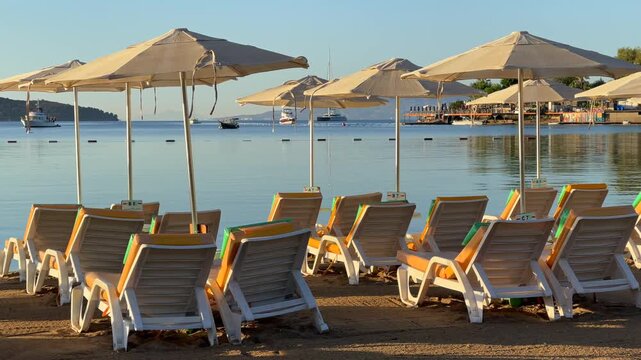 An empty sandy beach in Bitez, Bodrum, lined with neat rows of sun loungers and matching white umbrellas facing a calm, glassy blue bay beneath a bright morning sky.