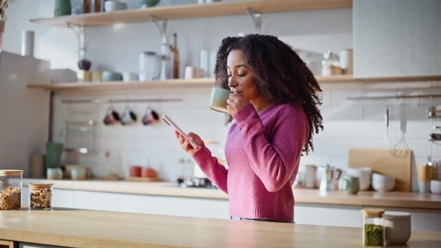 African woman sipping coffee holding phone at kitchen closeup. Smiling brunette