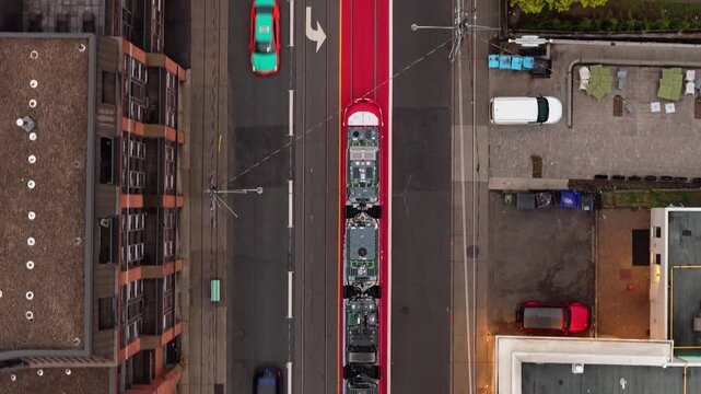 Top down aerial of a streetcar moving along a dedicated red right of way lane, cars driving beside buildings and a park in North America