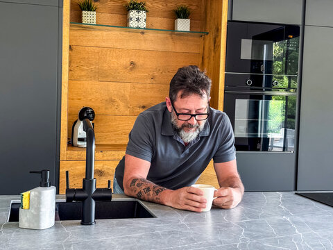 Bearded man with tattoo enjoying coffee in modern kitchen leaning on marble island