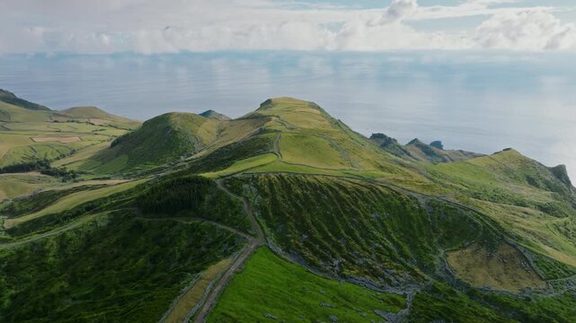 Aerial drone wide panorama of rolling green hills and patchwork fields stretching toward the calm Atlantic Ocean on Flores Island, Azores.